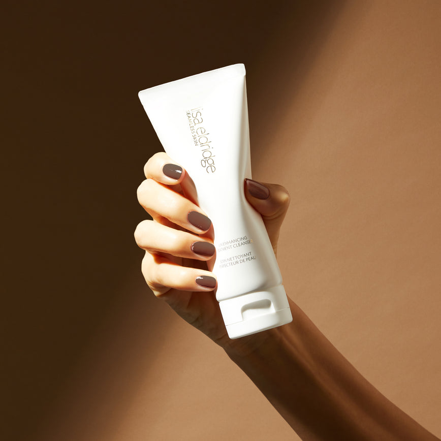 Model hand with brown nail narnish, holding Seamless Skin Enhancing Treatment Cleanser in front of a duo-toned tan background