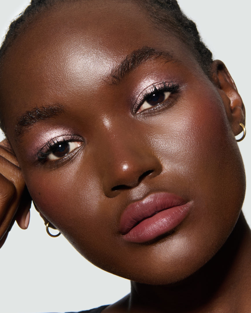 Close-up of a model's face against a neutral background, wearing Rosalie Liquid Lurex Liquid Eyeshadow, a warm, peachy rose.