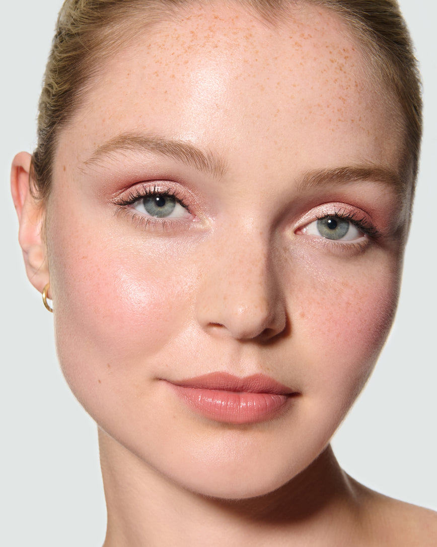 Close-up of a model's face against a neutral background, wearing Rosalie Liquid Lurex Liquid Eyeshadow, a warm, peachy rose.