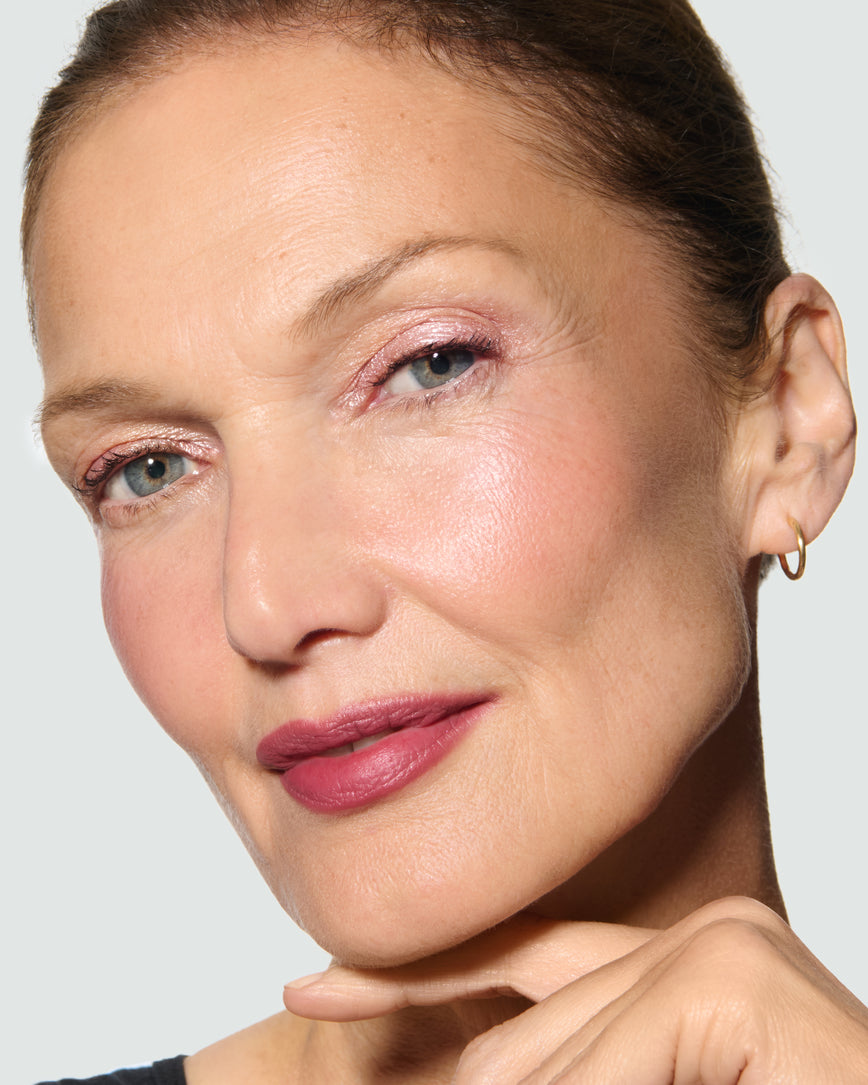 Close-up of a model's face against a neutral background, wearing Rosalie Liquid Lurex Liquid Eyeshadow, a warm, peachy rose.