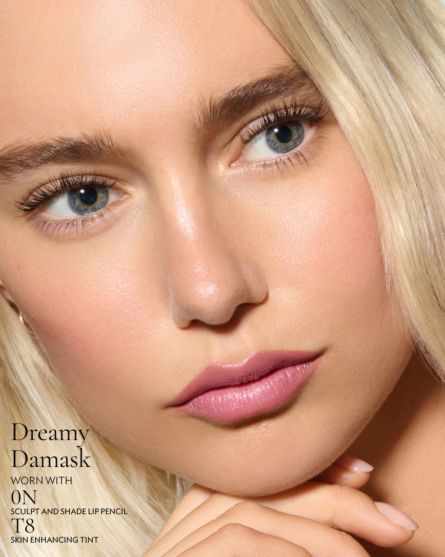 Close-up of a model's face against a neutral background, wearing Dreamy Damask Rouge Experience, Sculpt and Shade Lip Pencil in 0N and T8 Skin Enhancing Tint