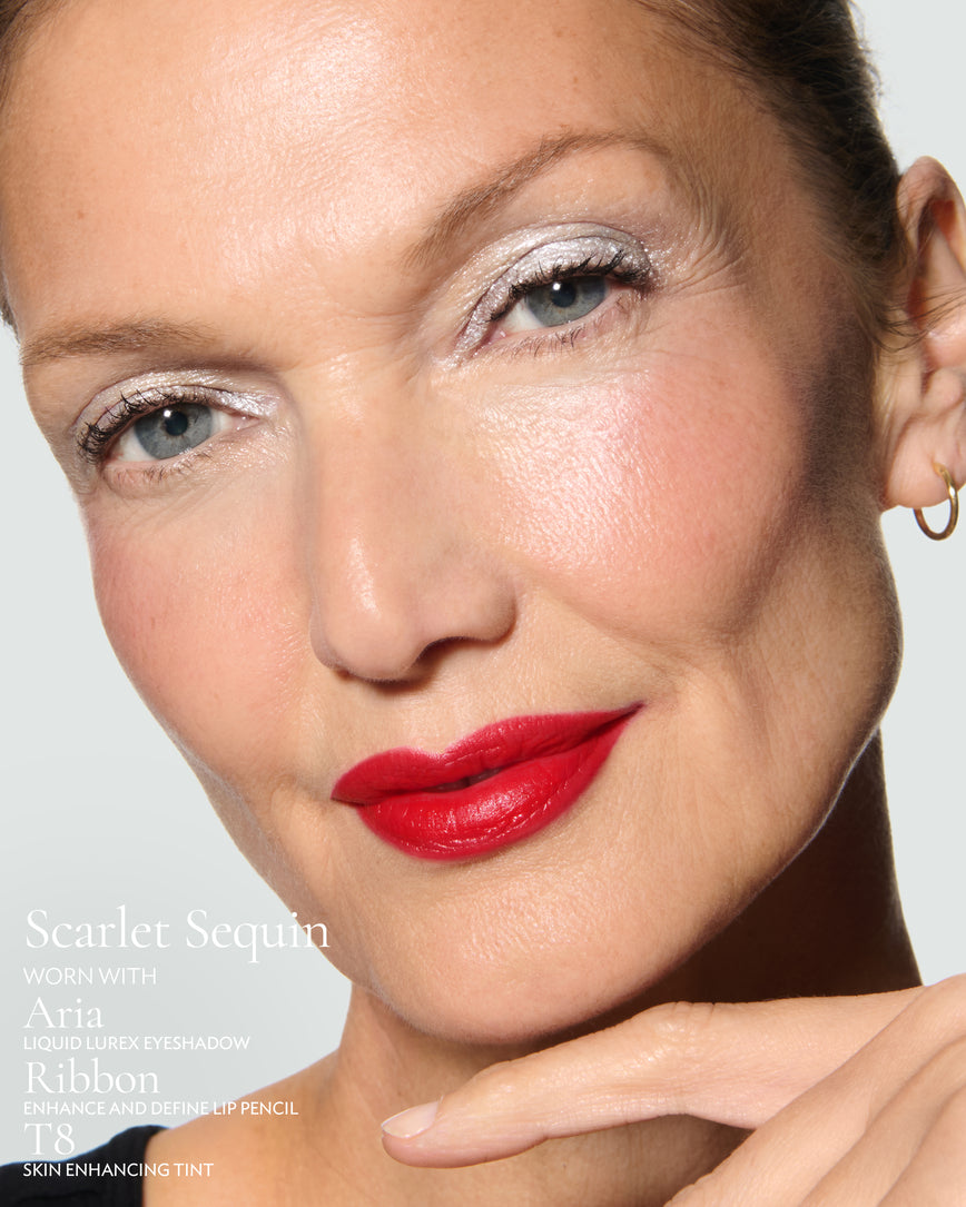 Close-up of a model's face against a neutral background, wearing Scarlet Sequin Rouge Experience, Aria Liquid Lurex, Ribbon Enhance and Define, and T8 Skin Enhancing Tint