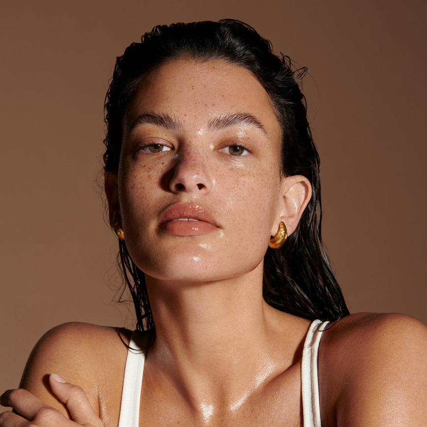 Portrait of a model with tan skin, glistening from the use of the Seamless Skin Enhancing Treatment Cleanser. Their dark hair is wet and pushed back from their face and goes down their back. On a brown background.