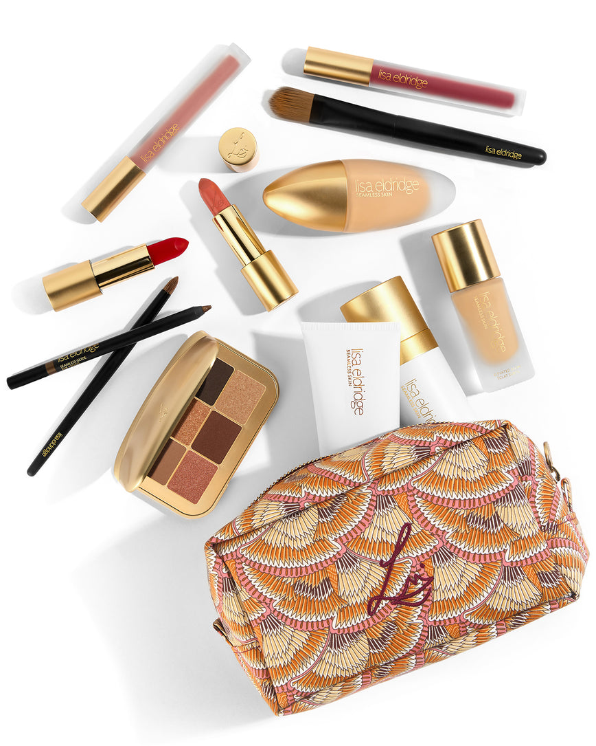 A beauty bag with Liberty print fabric which looks like overlapping fans that have brown, beige, orange, white and pink stripes sits on a white surface. Above it, as if spilled out from the bag, there is a collection of Lisa Eldridge products including foundations, lip products, pencils, a brush an eyeshadow palette (with neutral matte and shimmery colours) and a white tube and spray bottle that are half in the beauty bag and half out.