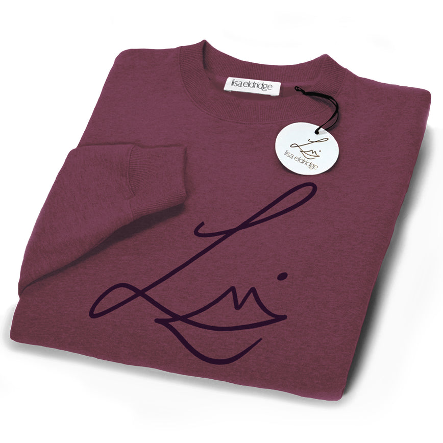 The Lisa Eldridge Studio Sweatshirt in velvet midnight folded up. The crewneck and the cuffs have a fine rib. The Lisa Eldridge logo is large on the front. There is a label that says “lisa eldridge’ sewn on the inside. Attached to the size label is a circular card tag on a black ribbon with the Lisa Eldridge logo in gold